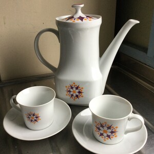 Może przedstawiać: Biały ceramiczny czajnik z kwiatowym wzorem w kolorze fioletowym i pomarańczowym oraz dwie pasujące filiżanki i spodki.