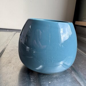 Könnte beinhalten: Ein blaugrüner Keramiktopf mit abgerundeter, leicht bauchiger Form. Der Topf hat eine glatte, glänzende Oberfläche.