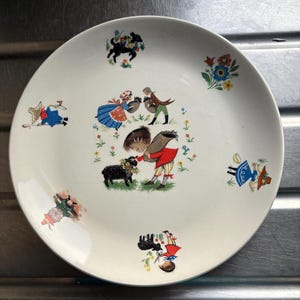 May include: A white ceramic plate with a colorful illustration of children and animals. The plate features a girl in a red dress petting a black sheep, a boy in a blue shirt holding a basket, and a girl in a blue dress holding a basket. There are also flowers and a black dog on the plate.