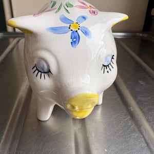 May include: White ceramic piggy bank with a whimsical design. The bank features a pig face with closed eyes, long eyelashes, and a yellow snout. It is decorated with blue and pink floral accents and yellow ears.