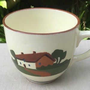 May include: A white ceramic mug with a brown rim and a painted scene of a house with a red roof and green trees.