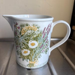 Può includere: Brocca in ceramica bianca con un design a bouquet floreale. Il bouquet presenta margherite, lavanda e altri fiori di campo nei toni del giallo, viola e verde. La brocca ha un manico e un beccuccio.