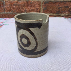 Pode incluir: Um pequeno jarro de cerâmica bege com um design de espiral marrom. O jarro tem um bico e uma alça.
