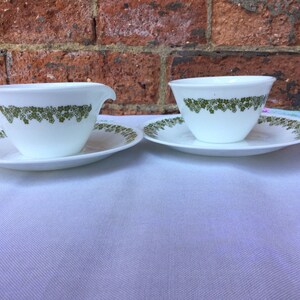 Puede incluir: Tazas y platillos blancos con un patrón floral verde. Una taza tiene asa y pico vertedor. Las tazas y platillos están sobre un mantel blanco. El fondo es una pared de ladrillo.