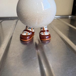 Puede incluir: Una maceta de cerámica blanca con efecto craquelado, apoyada sobre dos zapatos marrones y naranjas con detalles coloridos. La maceta tiene un cuerpo redondo y está diseñada para parecerse a una figura caprichosa.