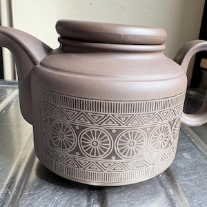 Puede incluir: Una tetera de cerámica marrón con un patrón geométrico. La tetera tiene una tapa y un asa.