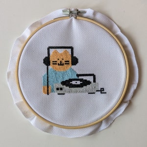 May include: Cross-stitch embroidery of a cat DJ wearing headphones, with a turntable. The cat is orange with black headphones and a light blue shirt. The turntable is gray and black, set in a wooden embroidery hoop.