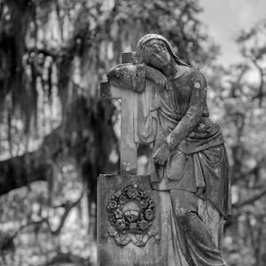 May include: A black and white photograph of a stone sculpture of a woman leaning against a cross. The woman is dressed in a long robe and has her head resting on her hand. The sculpture is weathered and has a detailed design.