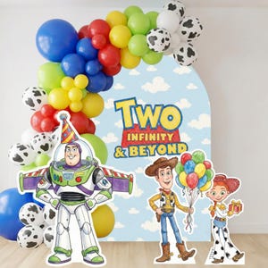 May include: A colorful party decoration featuring a balloon arch in blue, red, yellow, green, and white cow-print balloons. Cardboard cutouts of Buzz Lightyear, Woody, and Jessie stand in front of a backdrop with the text "Two Infinity & Beyond."