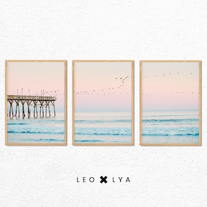 Puede incluir: Un conjunto de tres impresiones enmarcadas que muestran un paisaje marítimo con un muelle de madera, un océano azul y un cielo rosa y azul con una bandada de pájaros volando por encima.