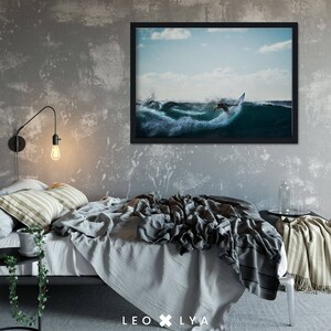 May include: Black framed print of a surfer riding a wave. The image is set against a blue sky with white clouds. The bed is covered with gray linens and a striped blanket. A black side table with a plant and a wall-mounted lamp with a lightbulb are also visible.