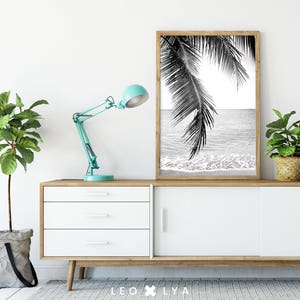 May include: Black and white photograph of palm tree leaves over a beach scene. The photo is framed and hanging on a white wall above a white dresser with three drawers.