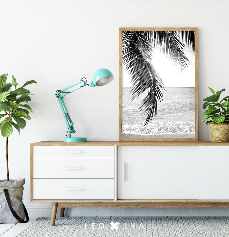 May include: Black and white photograph of palm tree leaves over a beach scene. The photo is framed and hanging on a white wall above a white dresser with three drawers.