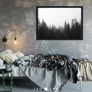 May include: A black and white photograph of a forest with tall trees, framed in a black frame, hangs on a grey wall above a bed with a grey and white striped throw blanket and a grey duvet cover.