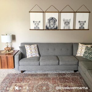 May include: A gray sectional sofa with two throw pillows, one with a geometric pattern in shades of red, white, and yellow, and the other with a geometric pattern in shades of green and white. The sofa is in front of a wall with four framed black and white prints of lion faces. The wall is a light beige color. A small wooden end table with a lamp is to the left of the sofa. A red and cream patterned rug is on the floor in front of the sofa. The text "@loveluxemmama" is on the rug.