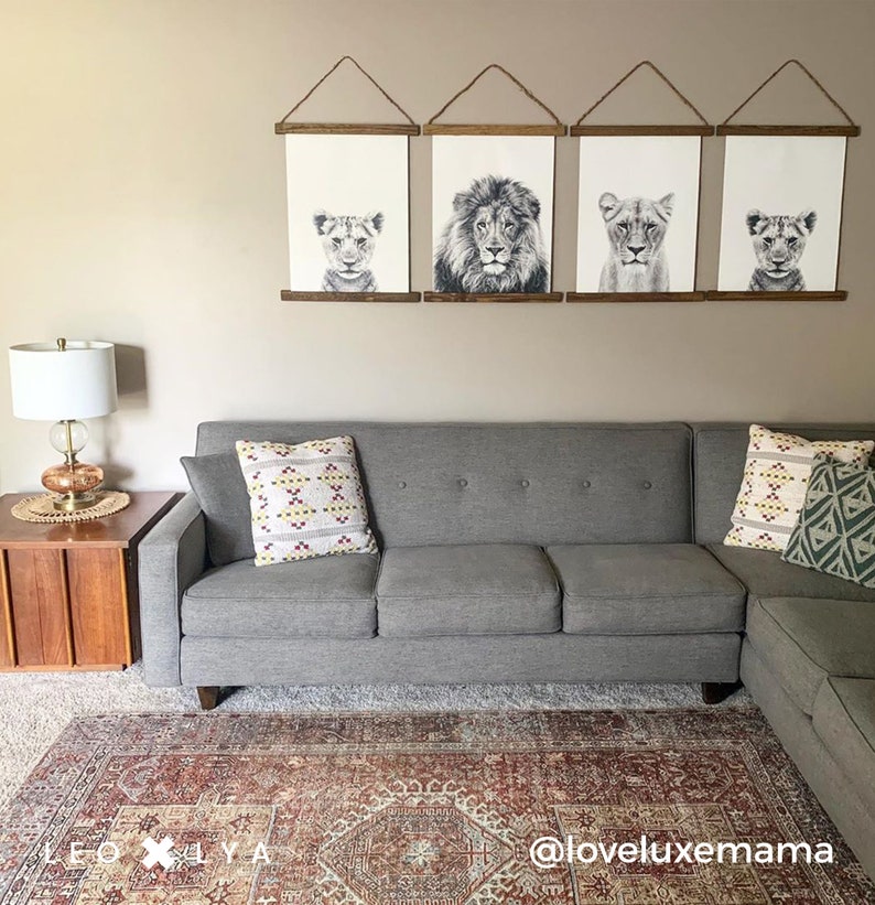 May include: A gray sectional sofa with two throw pillows, one with a geometric pattern in shades of red, white, and yellow, and the other with a geometric pattern in shades of green and white. The sofa is in front of a wall with four framed black and white prints of lion faces. The wall is a light beige color. A small wooden end table with a lamp is to the left of the sofa. A red and cream patterned rug is on the floor in front of the sofa. The text "@loveluxemmama" is on the rug.