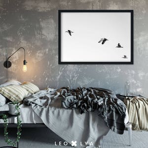 May include: A black framed print of a minimalist photograph of five birds flying in formation against a white background. The print is hanging on a gray wall above a bed with a gray and white striped throw blanket.