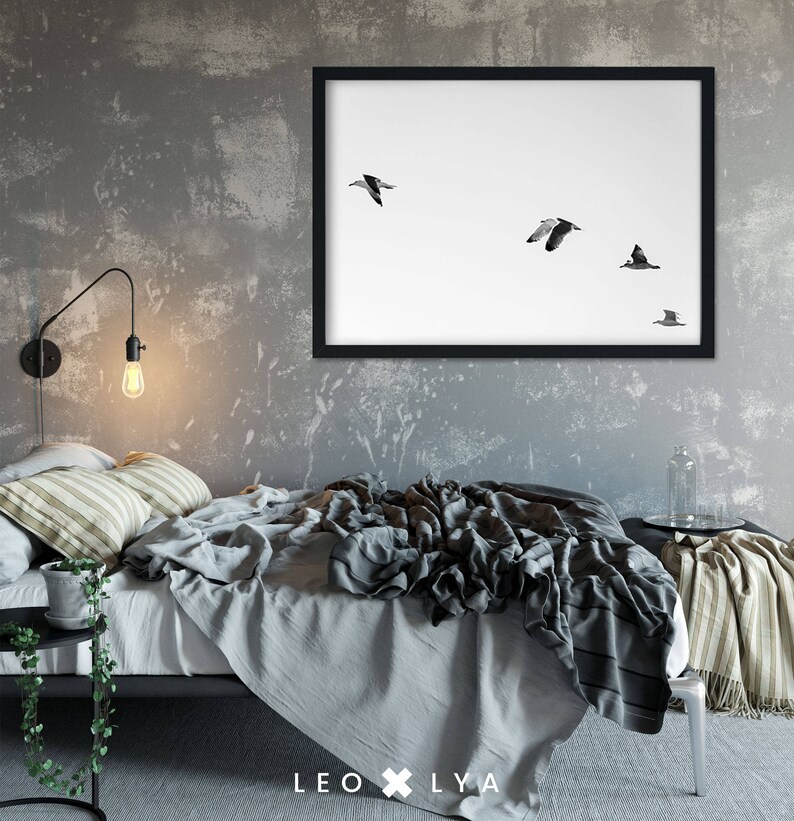 May include: A black framed print of a minimalist photograph of five birds flying in formation against a white background. The print is hanging on a gray wall above a bed with a gray and white striped throw blanket.