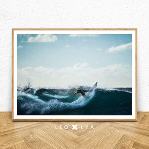 May include: A framed print featuring a surfer riding a wave. The image captures the surfer mid-action, with the wave cresting and breaking. The sky is a light blue with scattered clouds. The print is in a wooden frame, with the text "LEO X LYA" at the bottom.