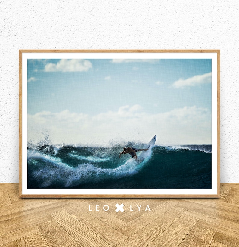 May include: A framed print featuring a surfer riding a wave. The image captures the surfer mid-action, with the wave cresting and breaking. The sky is a light blue with scattered clouds. The print is in a wooden frame, with the text "LEO X LYA" at the bottom.