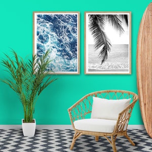 May include: Two black and white framed prints of ocean waves and palm trees hang on a turquoise wall. A white wicker chair with a white cushion sits in front of the prints. A wooden surfboard leans against the wall to the right of the prints.