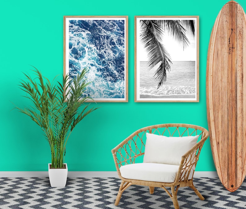May include: Two black and white framed prints of ocean waves and palm trees hang on a turquoise wall. A white wicker chair with a white cushion sits in front of the prints. A wooden surfboard leans against the wall to the right of the prints.