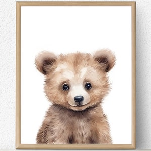 May include: A framed watercolor print featuring a cute brown bear cub. The bear has soft, fluffy fur, a small black nose, and big, dark eyes. The artwork is set against a white background and framed in a light wood frame, suitable for nursery decor.