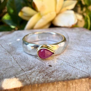 May include: A silver ring featuring a hammered texture and a single pear-shaped, deep pink gemstone set in a gold-toned bezel. The band has a polished finish and a rounded profile, showcasing a contrast between the silver metal and the vibrant stone.