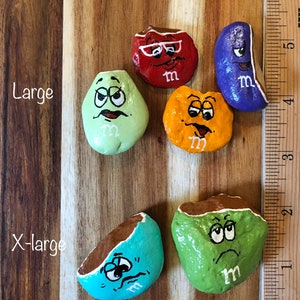 May include: Six painted rocks with different expressions and colors, resembling M&M's candies. The rocks are painted in red, green, blue, orange, and brown. The rocks are arranged on a wooden surface with a ruler in the background. The rocks are labeled as "Large" and "X-Large".