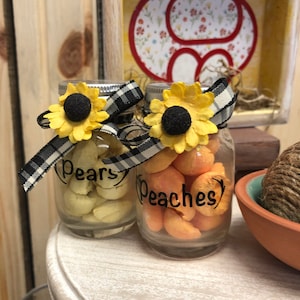 May include: Two glass jars with black and white checkered ribbon and yellow sunflower accents. The jars contain yellow and orange candies. The jars are labeled "Pears" and "Peaches".