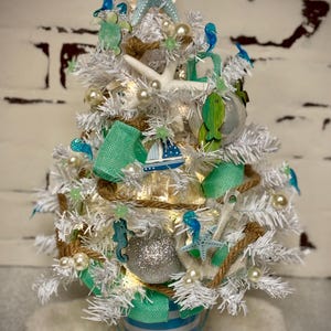 May include: A small, white Christmas tree decorated with a nautical theme. It features blue starfish, seahorses, and a sailboat, along with pearls and teal ribbon. The tree sits in a blue and white striped bucket, surrounded by seashells.