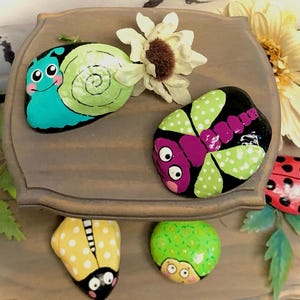 May include: Hand-painted rocks featuring whimsical insect designs. A teal snail, a black and green butterfly, a red ladybug, a yellow ladybug, and a green bug are displayed on a wooden surface. Decorative flowers add a natural touch.