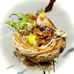 May include: A small bird figurine sits in a brown twig nest with a green egg and a brown nut. The nest is decorated with green leaves and yellow flowers.