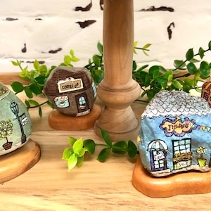 May include: Four painted rock houses with different storefronts, including a bakery, coffee shop, flower shop, and a cafe. Each rock house is sitting on a wooden base.