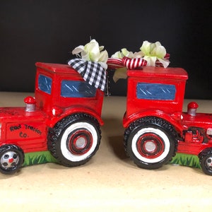 May include: Two red toy tractors with white tires and black accents. The tractors are decorated with a white and black checkered fabric and white flowers. The front tractor has the words "Red Tractor Co." printed on the side.