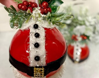 May include: Red and white Christmas ornament with a Santa Claus design. The ornament has a black belt, white trim, and black buttons. It is decorated with green holly and red berries.