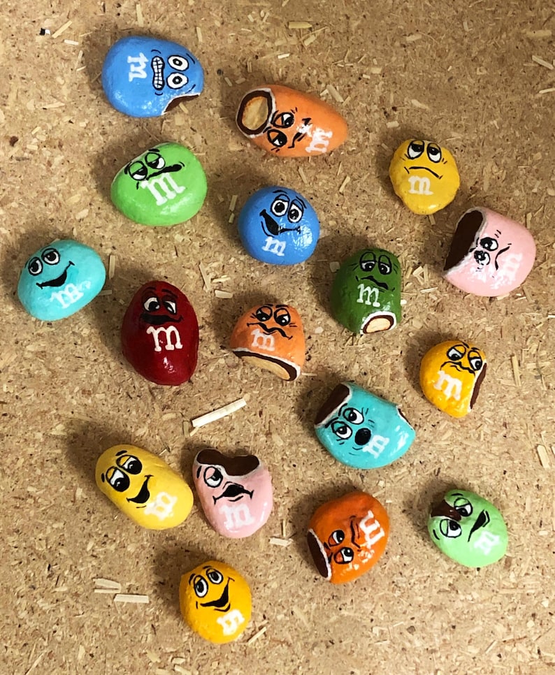 May include: A collection of 15 painted rocks, each depicting a different M&M's character with various expressions. The rocks are painted in a variety of colors, including red, blue, green, yellow, orange, and pink.