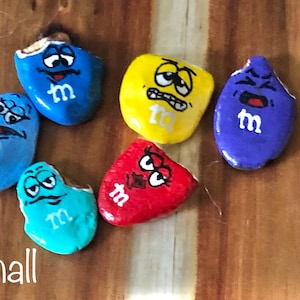 May include: Six painted rocks shaped like M&M's candies with different facial expressions. The rocks are painted in blue, yellow, purple, red, and turquoise. The word "Small" is written on the wooden surface below the rocks.