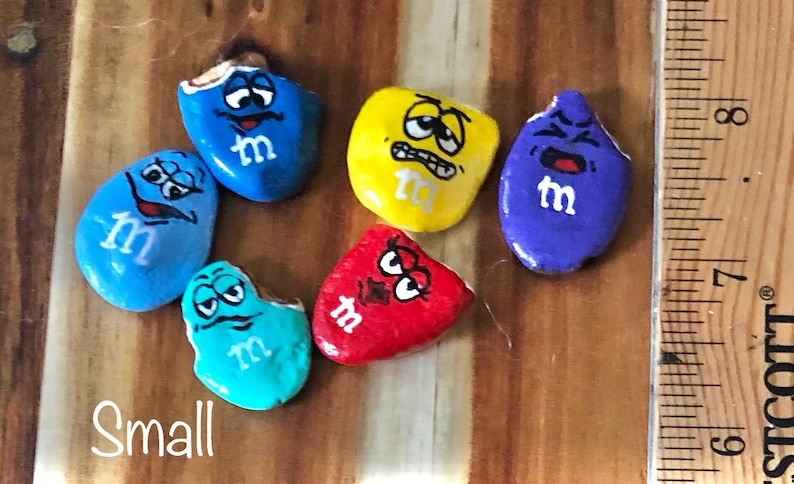 May include: Six painted rocks shaped like M&M's candies with different facial expressions. The rocks are painted in blue, yellow, purple, red, and turquoise. The word "Small" is written on the wooden surface below the rocks.