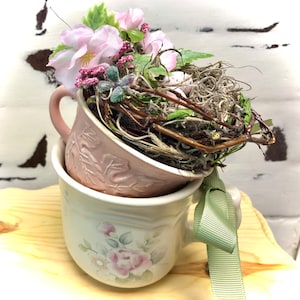 May include: Two pink teacups with floral designs, one nested inside the other. The top cup is decorated with a nest of twigs and faux pink flowers.