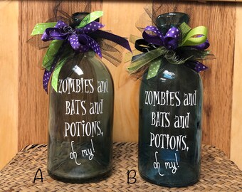 May include: Two glass bottles with a black, green, and purple ribbon bow. The bottles are decorated with the text "ZOMBIES and BATS and POTIONS, oh my!"