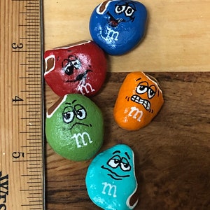 May include: Five painted rocks with different expressions, each painted to look like an M&M candy. The rocks are painted in blue, red, green, orange, and turquoise. Each rock has a white "m" painted on it.