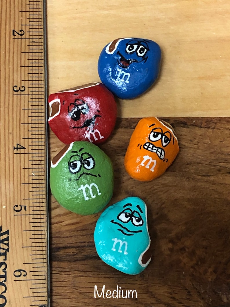 May include: Five painted rocks with different expressions, each painted to look like an M&M candy. The rocks are painted in blue, red, green, orange, and turquoise. Each rock has a white "m" painted on it.