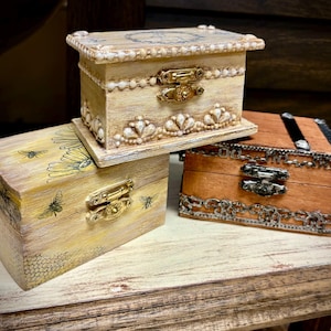 May include: Three decorative wooden boxes with different designs. The top box is white with a floral design and a gold latch. The middle box is yellow with a bee and flower design and a gold latch. The bottom box is brown with a metal latch and a decorative border.