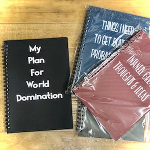 May include: Three black spiral bound notebooks with white text. The first notebook says "My Plan For World Domination". The second notebook says "Things I Need To Get Done Probably". The third notebook says "Manly Great Thoughts & Ideas".