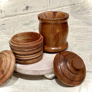 May include: Three wooden containers with lids. The containers are made of different types of wood and have different patterns. The containers are sitting on a white surface.