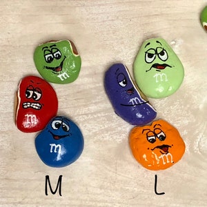 May include: Nine painted rocks in various sizes and colors, each depicting a cartoon M&M candy with a different facial expression. The rocks are labeled with the sizes S, M, L, and XL.