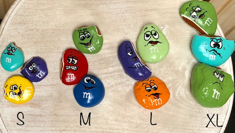May include: Nine painted rocks in various sizes and colors, each depicting a cartoon M&M candy with a different facial expression. The rocks are labeled with the sizes S, M, L, and XL.
