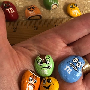 May include: A collection of hand-painted rock M&M's. The rocks are painted to look like the popular candy with different expressions. The rocks are various colors, including green, blue, orange, yellow, and brown.