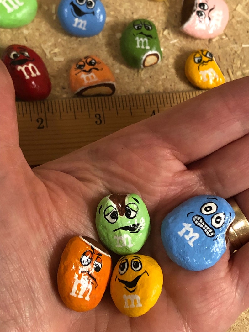 May include: A collection of hand-painted rock M&M's. The rocks are painted to look like the popular candy with different expressions. The rocks are various colors, including green, blue, orange, yellow, and brown.